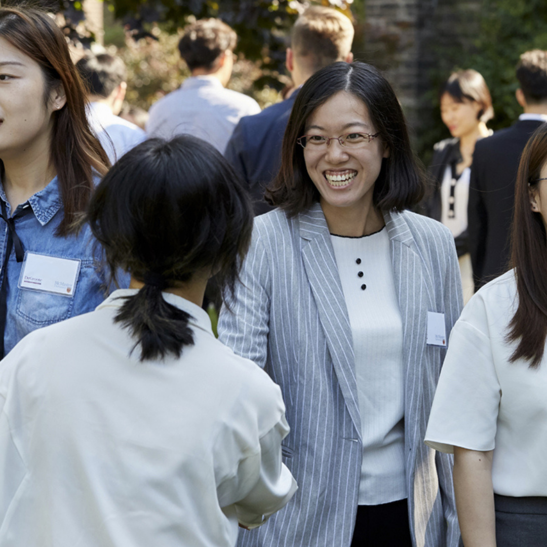 Two women smiling and shaking hands in a crowd of other students.