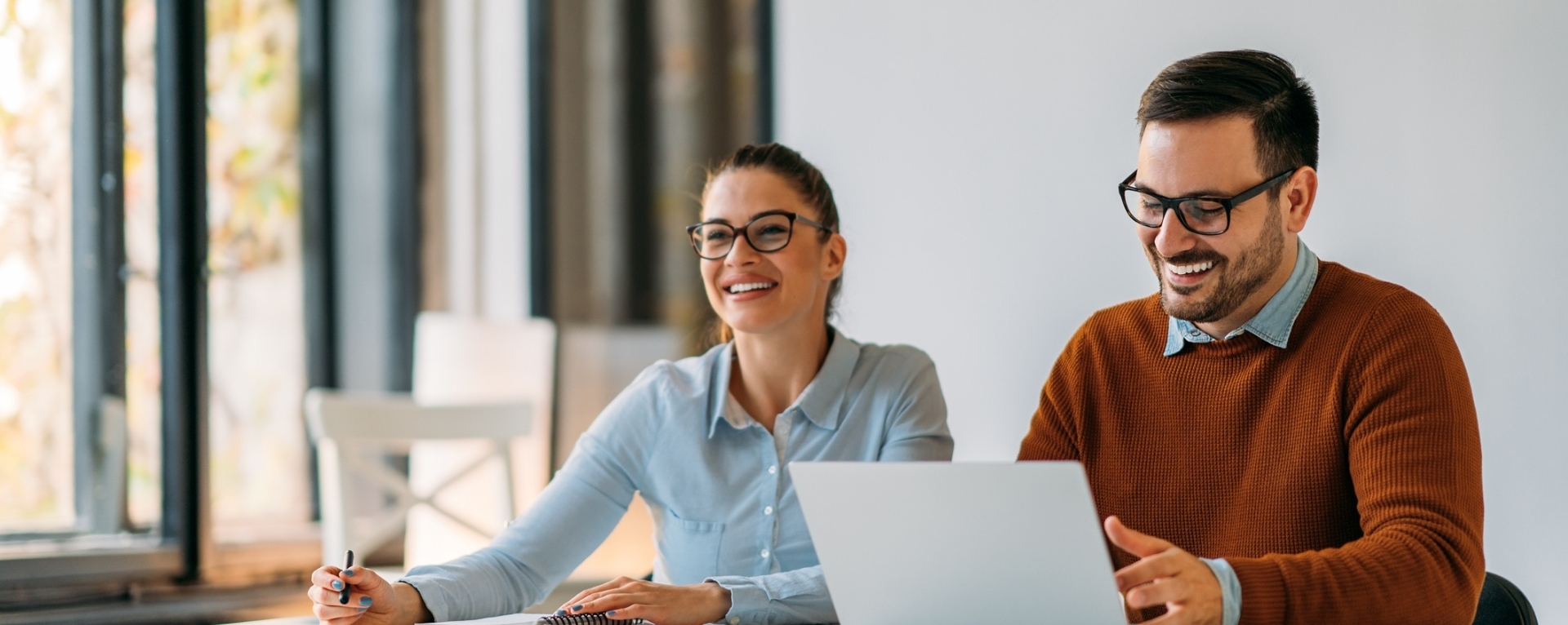 A man and woman seated in a office smiling and taking notes on laptop and notepad respectively.